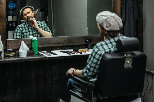 homem sentado na cadeira de barbeiro em frente ao espelho e tocando sua barba