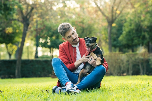Homem sentado na grama acariciando um cachorro sentado no colo dela em um parque