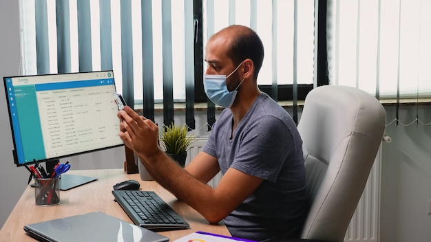 Homem sentado na sala de escritório na mesa, digitando no telefone, usando máscara de proteção. Freelancer trabalhando em um novo espaço de trabalho normal, conversando, falando, escrevendo, usando telefone celular com tecnologia de internet