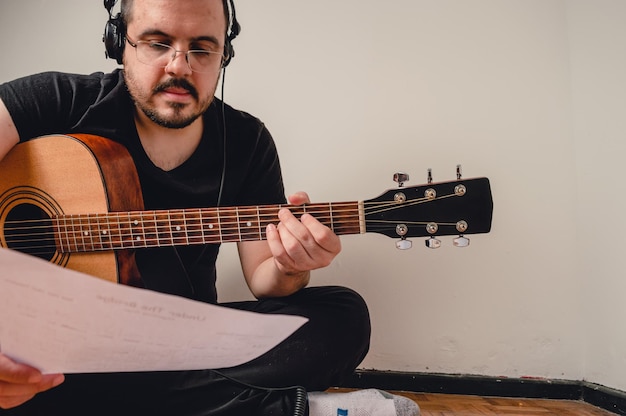 Homem sentado na sala de sua casa praticando violão fazendo um acorde lendo partituras