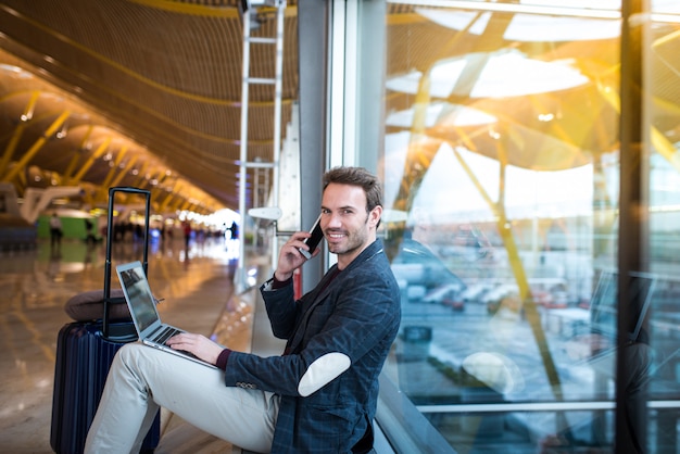 Homem sentado no aeroporto usando o laptop e telefone celular ao lado da janela