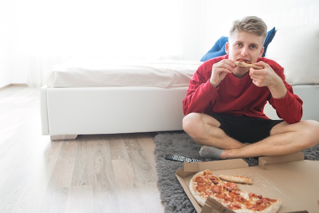 Foto homem sentado no chão à beira da cama com uma caixa de pizza e um pedaço nas mãos