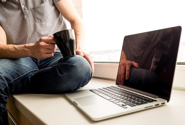 Homem sentado no peitoril da janela com café e laptop