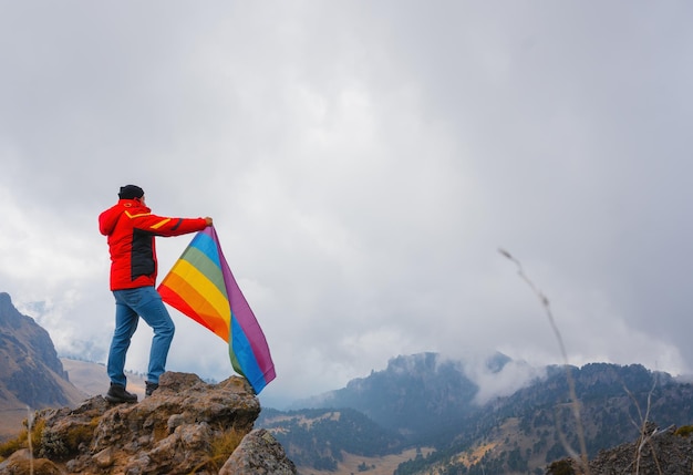 Homem sentado no topo de uma montanha ergueu a bandeira do símbolo LGBT do arco-íris para o céu ensolarado