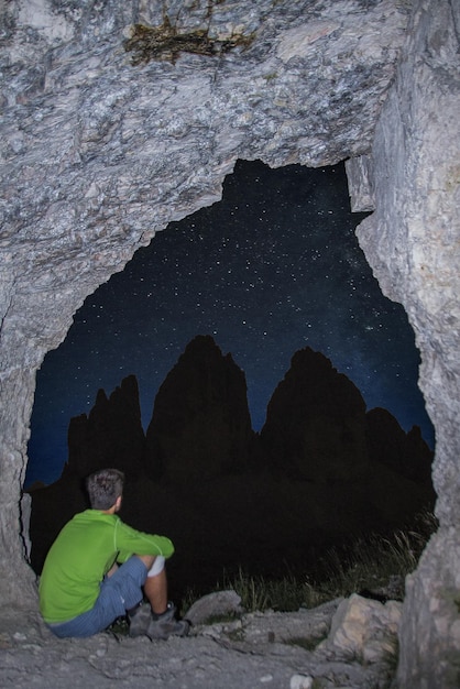 Foto homem sentado numa caverna a olhar para o céu estrelado