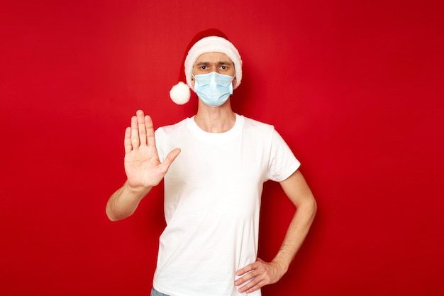 Homem sério com máscara médica, boné de ano novo mostrando o sinal de pare com a mão estendida à sua frente