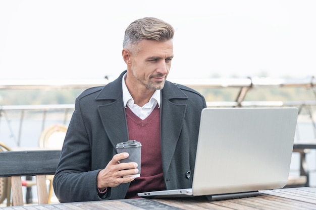 Homem sério de diretor trabalhando online no laptop tomando café no café ao ar livre