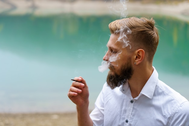 Homem sério e bonito, jovem pensativo, fuma sistema de produtos de tabaco de aquecimento ao ar livre no bac ...