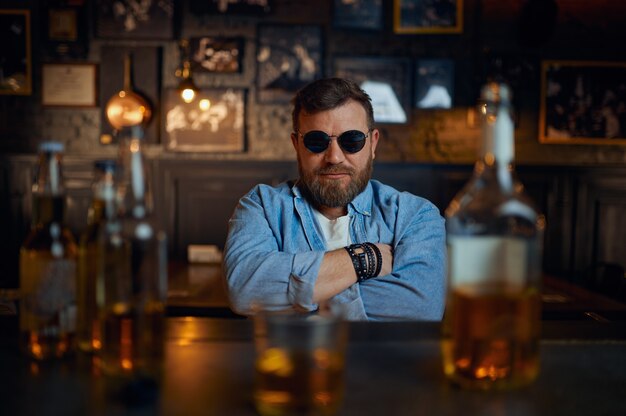 Homem sério em óculos de sol, sentado no balcão do bar. Um homem descansando em um bar, emoções humanas, atividades de lazer, vida noturna