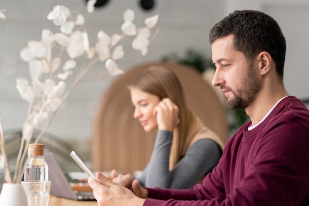 Homem sério em roupa casual rolando no touchpad enquanto está sentado no café com uma jovem