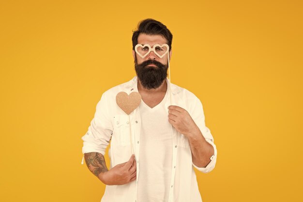 Homem sério hipster em óculos de festa homem barbudo hipster fundo amarelo coração de amor para o dia de São Valentim feriado feliz aniversário homem brutal com barba se apaixonar data romântica