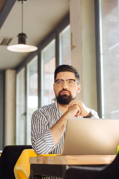 Homem sério. homem bonito e bonito sentado em frente ao laptop enquanto pensa em seu trabalho