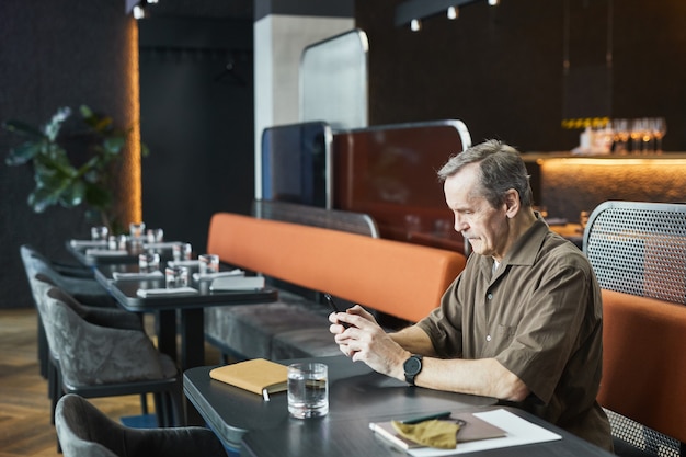 Homem sério sênior com camisa marrom e relógio sentado à mesa e mandando mensagem de texto no telefone enquanto espera no café