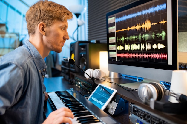 Foto homem sério tocando uma das teclas do piano enquanto olha as formas de onda do som na tela do computador em frente