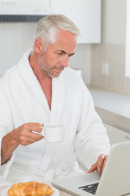 Homem serio usando o laptop no café da manhã em um roupão de banho