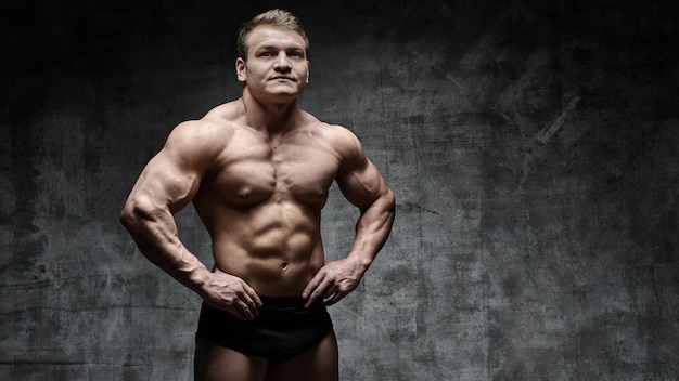 Homem sexy fisiculturista posando em fundo escuro em shorts pretos. Bonito corpo masculino bombeado isolado com espaço livre para publicidade