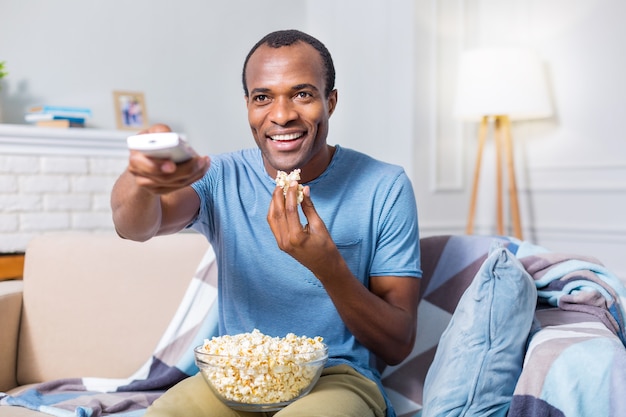 Homem simpático e feliz sorrindo e assistindo TV enquanto segura uma tigela com pipoca