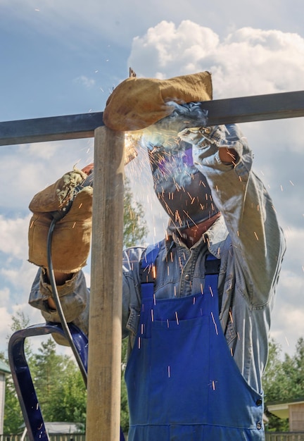 Homem soldador em uma máscara de soldagem, uniforme de construção e luvas de proteção cozinha metal em um canteiro de obras de rua. Construção de um pavilhão, pérgula perto de uma casa de campo em um dia de verão.