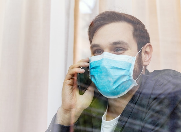 Homem solitário em quarentena com covid-19 em isulação, falando por telefone com a família, olha pela janela. Prevenção de pandemia de coronavírus. Homem em quarentena de máscara médica de proteção. Conceito de cuidados de saúde de medicina