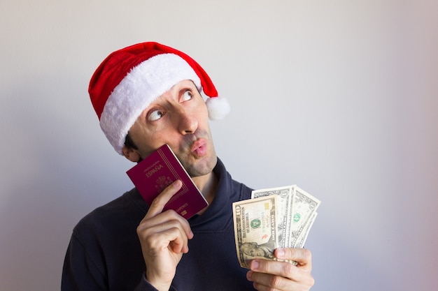 Homem sonhador com chapéu de Papai Noel segurando passaporte e notas de dólar com olhar pensativo nas férias de Natal