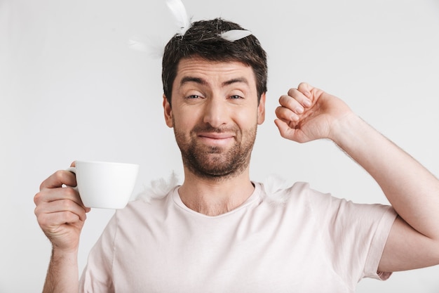 homem sonolento com cerdas em uma camiseta casual bebendo café enquanto fica em pé sob as penas que caem, isoladas sobre uma parede branca