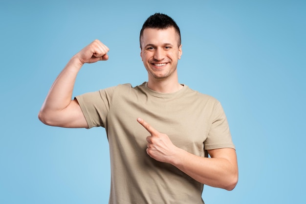 Homem sorridente atlético forte com camiseta pastel apontando bíceps mostrando força