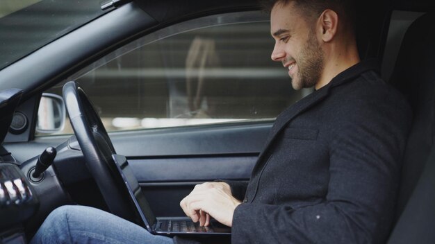 Homem sorridente atraente digitando laptop enquanto está sentado dentro de seu carro ao ar livre