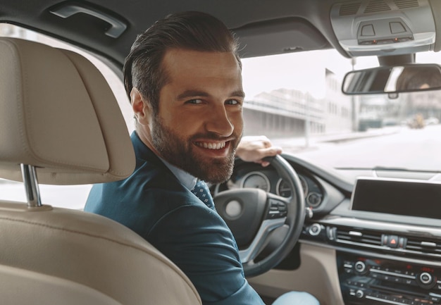 Homem sorridente barbudo com roupa formal posando em um carro