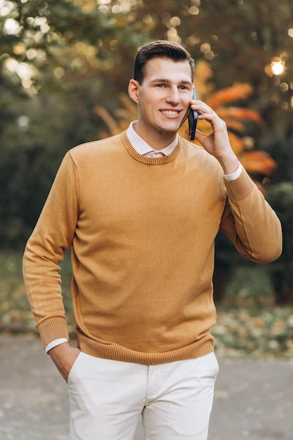 Homem sorridente bonito moderno em roupas amarelas e brancas falando ao telefone no parque ao pôr do sol