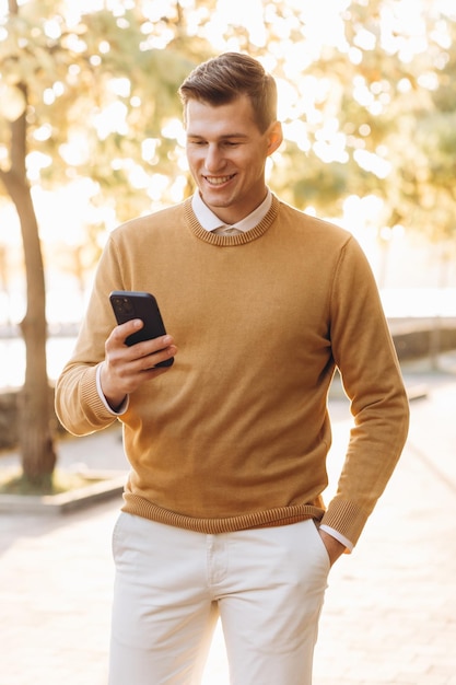 Homem sorridente bonito moderno em roupas amarelas e brancas falando ao telefone no parque ao pôr do sol