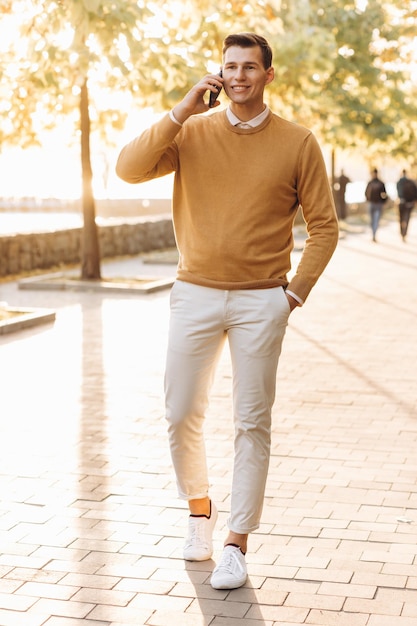 Homem sorridente bonito moderno em roupas amarelas e brancas falando ao telefone no parque ao pôr do sol