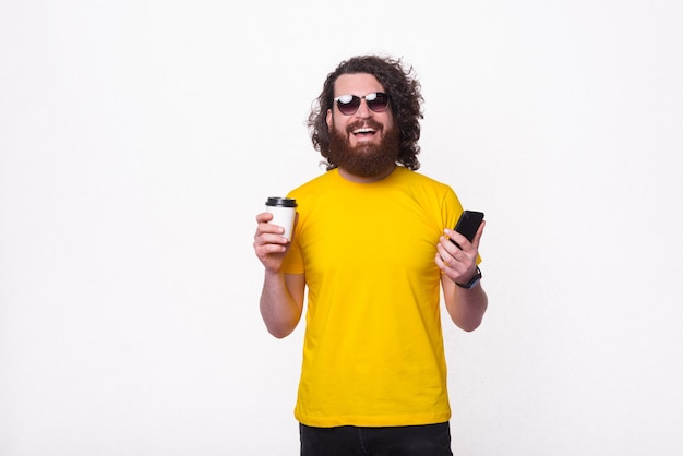 Homem sorridente com barba em uma camiseta amarela segurando uma xícara de café e um smartphone