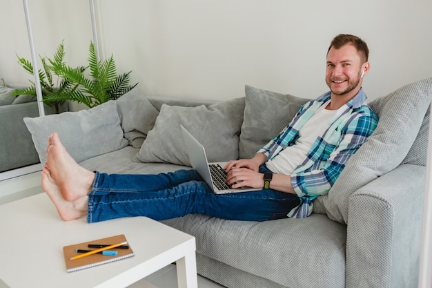 homem sorridente com camisa sentado relaxado no sofá em casa à mesa trabalhando online no laptop de casa