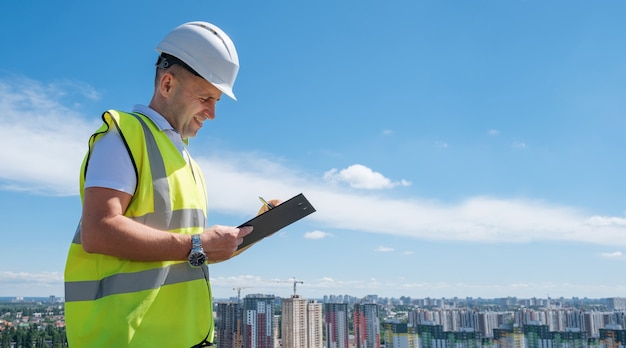 Homem sorridente com capacete branco no canteiro de obras escreve notas na prancheta