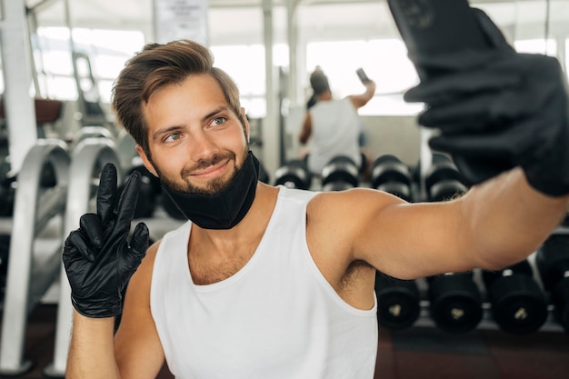 Foto homem sorridente com máscara médica tirando uma selfie na academia