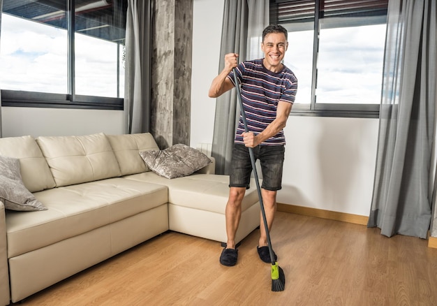 Homem sorridente com roupas casuais e chinelos varrendo sua sala de estar. Corpo todo.