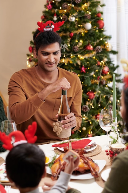 Homem sorridente com tiara de chifres de rena abrindo uma garrafa de vinho para os convidados no jantar de natal