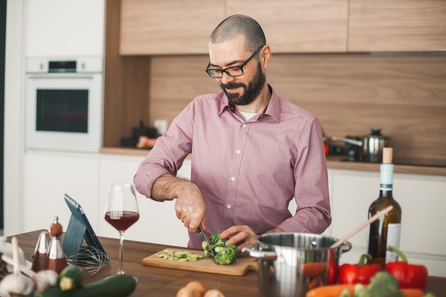 Homem sorridente com um copo de vinho, cozinhar ensopado de vegetais usando o tablet. Ele está cortando brócolis, abobrinha, pimenta vermelha, cebola e outros vegetais.