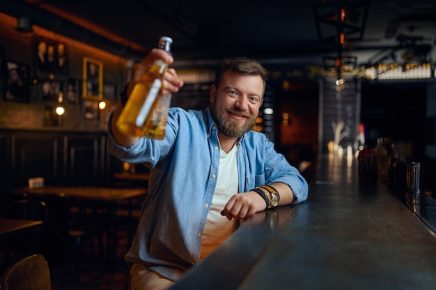 Homem sorridente com uma garrafa de cerveja no balcão do bar. Um homem descansando em um bar, emoções humanas, atividades de lazer, vida noturna