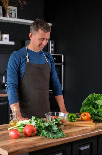 Homem sorridente cozinhando salada, cortando legumes frescos na cozinha preta moderna