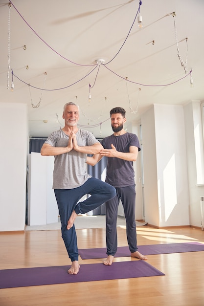 Homem sorridente de cabelos grisalhos com as palmas das mãos juntas no coração fazendo a pose da árvore