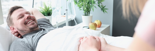 Homem sorridente deitado na cama do hospital, sua mão é segurada por uma mulher, apoiando entes queridos com doenças