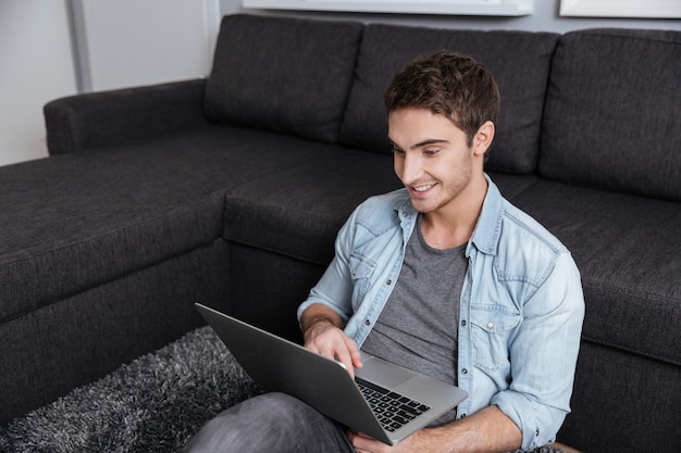 Homem sorridente e casual sentado no tapete com o laptop em casa