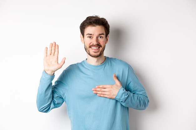 Homem sorridente e hospitaleiro erguendo o braço e segurando o coração, fazendo promessa dizer a verdade, jurar ou prestar juramento, de pé sobre um fundo branco