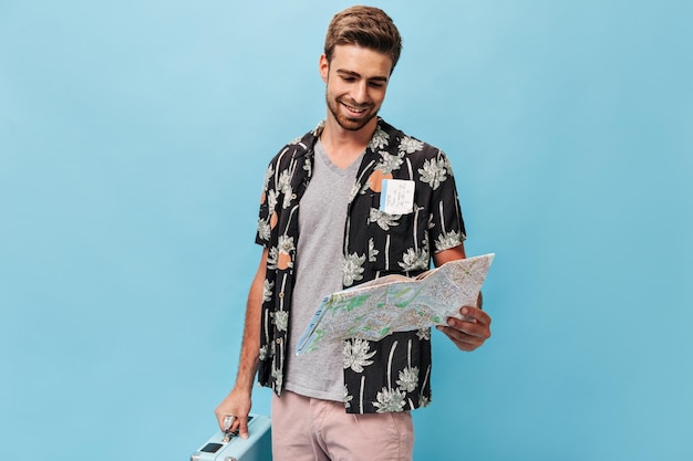 Homem sorridente elegante com barba em camisa de manga curta pintada e camiseta cinza olhando para o mapa em fundo azul isolado