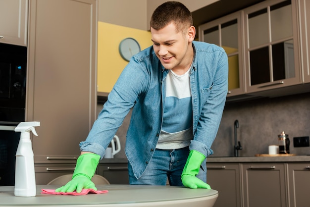 Homem sorridente em luvas de borracha, mesa de limpeza com pano e detergente na cozinha em casa