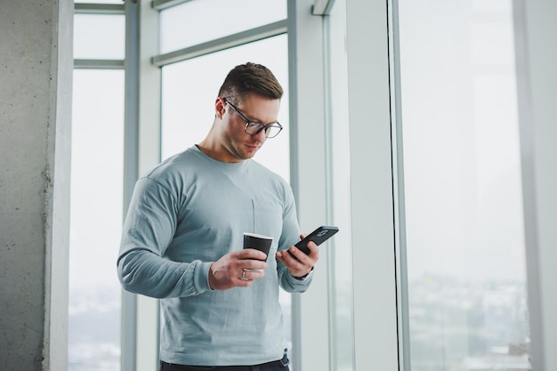 Homem sorridente em roupas casuais em pé perto da janela com café e olhando para smartphone no espaço de trabalho moderno com janela grande durante o dia