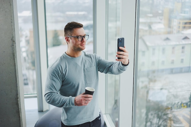 Homem sorridente em roupas casuais em pé perto da janela com café e olhando para smartphone no espaço de trabalho moderno com janela grande durante o dia