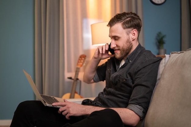 Homem sorridente está sentado na sala de estar no sofá com seu laptop nas pernas navegando férias