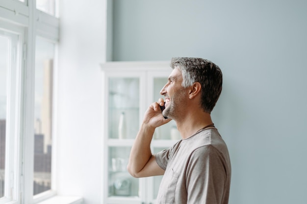 Homem sorridente falando em um smartphone em pé perto da janela